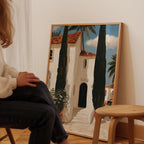 A person is sitting in front of a large painting depicting a white building with a red roof, surrounded by palm trees, in a room with a wooden stool nearby.