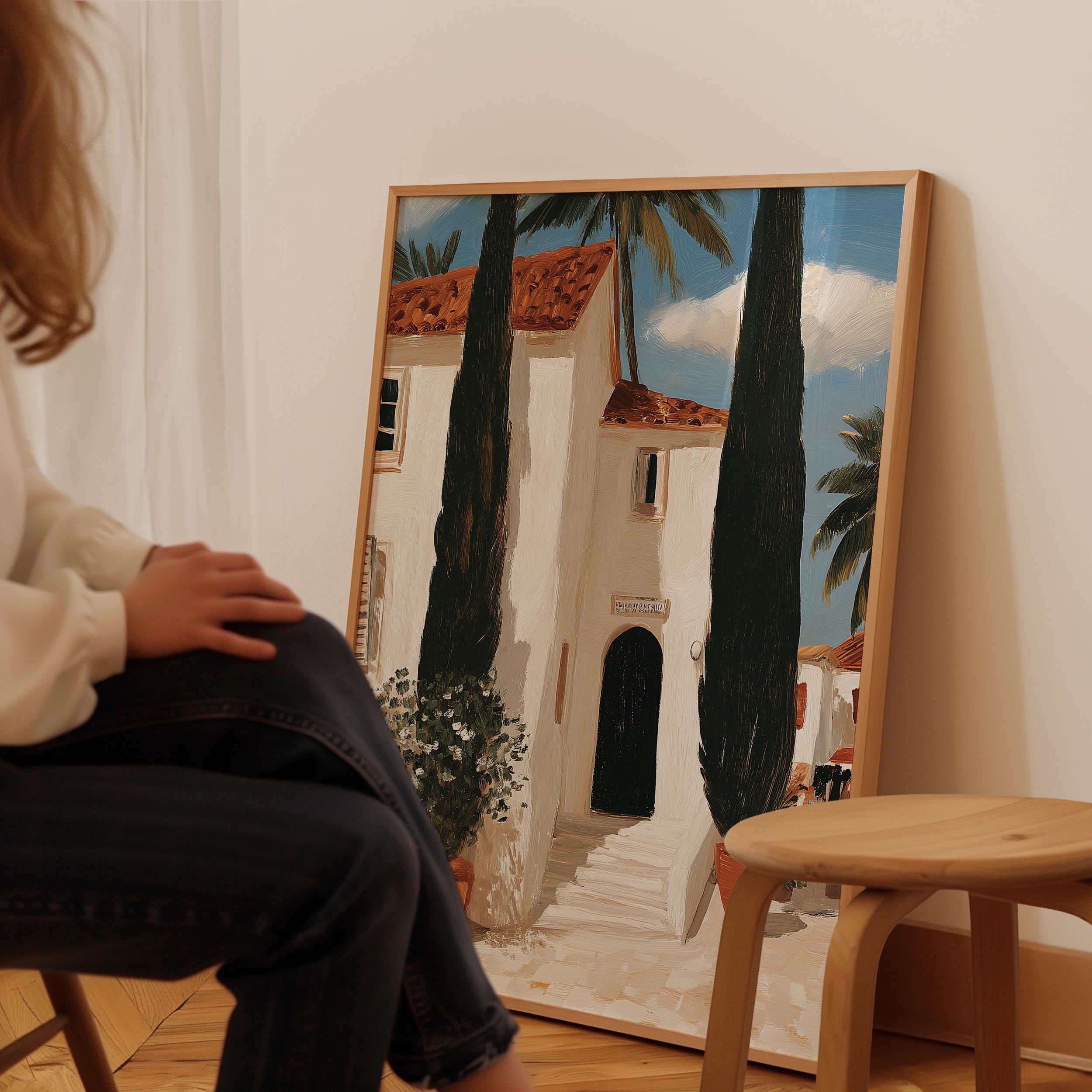 A person is sitting in front of a large painting depicting a white building with a red roof, surrounded by palm trees, in a room with a wooden stool nearby.