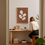 A woman is sitting at a wooden desk, surrounded by various objects, including a plant, books, and a framed picture on the wall.