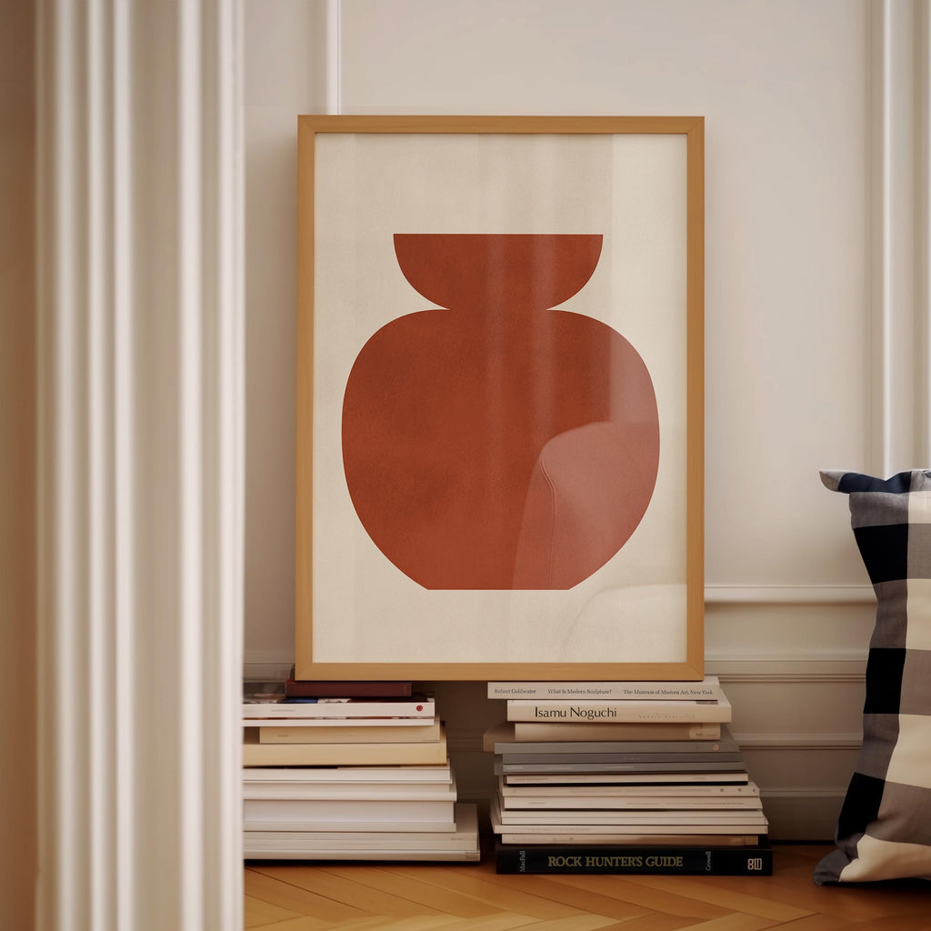 a framed artwork of a red vase on a white wall, surrounded by a stack of books and a pillow.