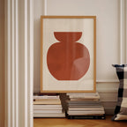 a framed artwork of a red vase on a white wall, surrounded by a stack of books and a pillow.