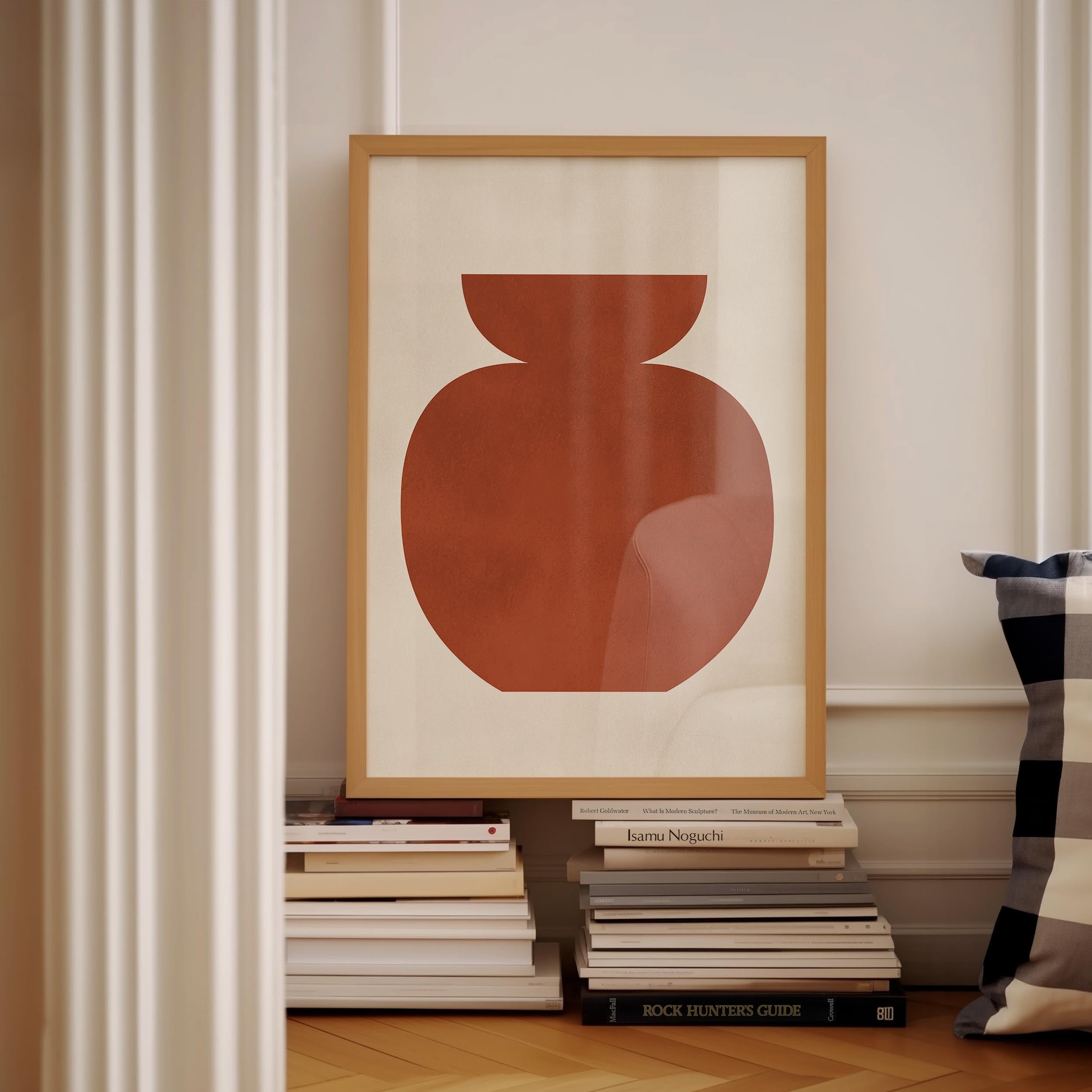 a framed artwork of a red vase on a white wall, surrounded by a stack of books and a pillow.