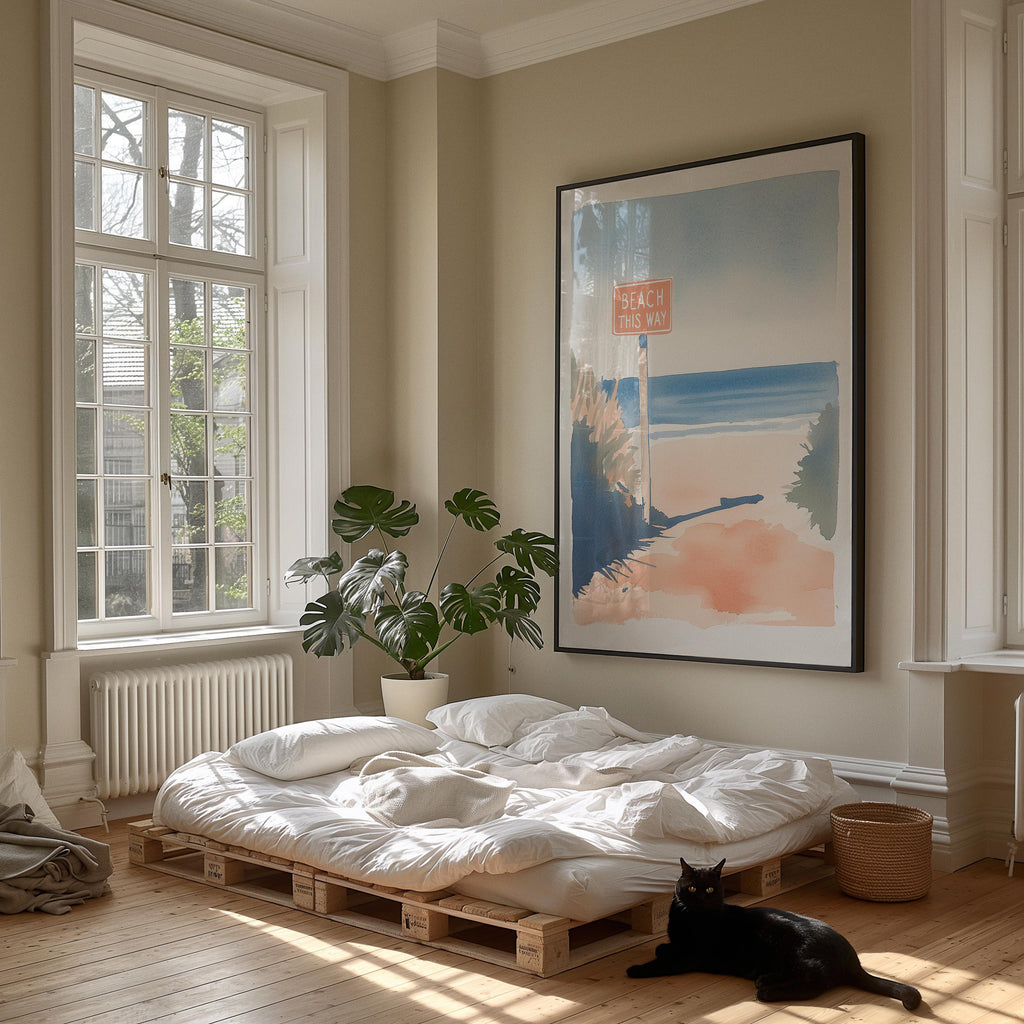 A cozy bedroom with a large window, a bed with white sheets, a potted plant, and a framed artwork depicting a beach scene. A black cat is sitting on the floor in front of the bed.