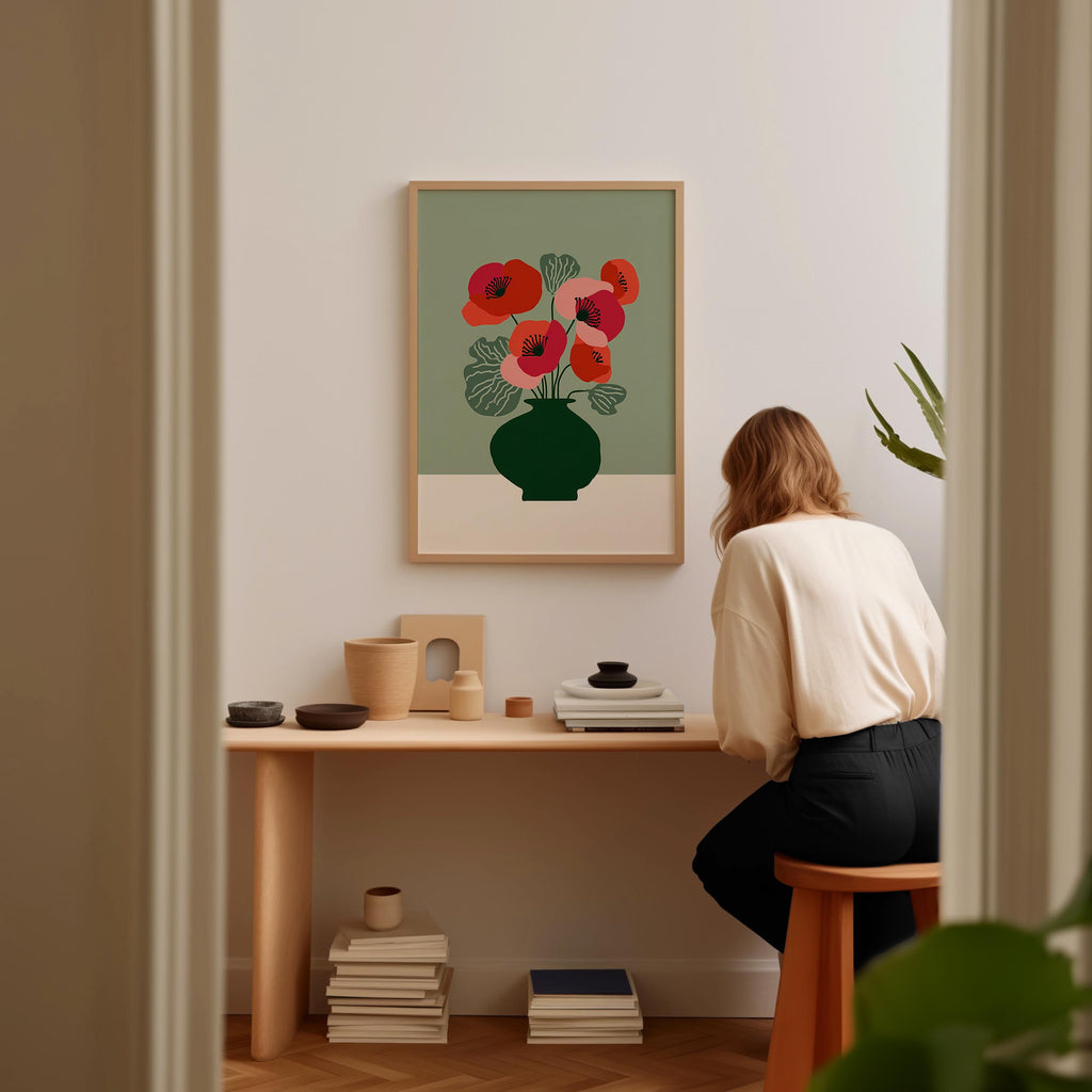 A woman is sitting at a wooden desk, looking at a framed print of a vase with red flowers on the wall behind her.