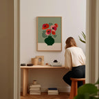 A woman is sitting at a wooden desk, looking at a framed print of a vase with red flowers on the wall behind her.