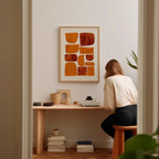 A woman is sitting at a wooden desk, surrounded by various objects, including a framed abstract artwork, a stack of books, and a plant.