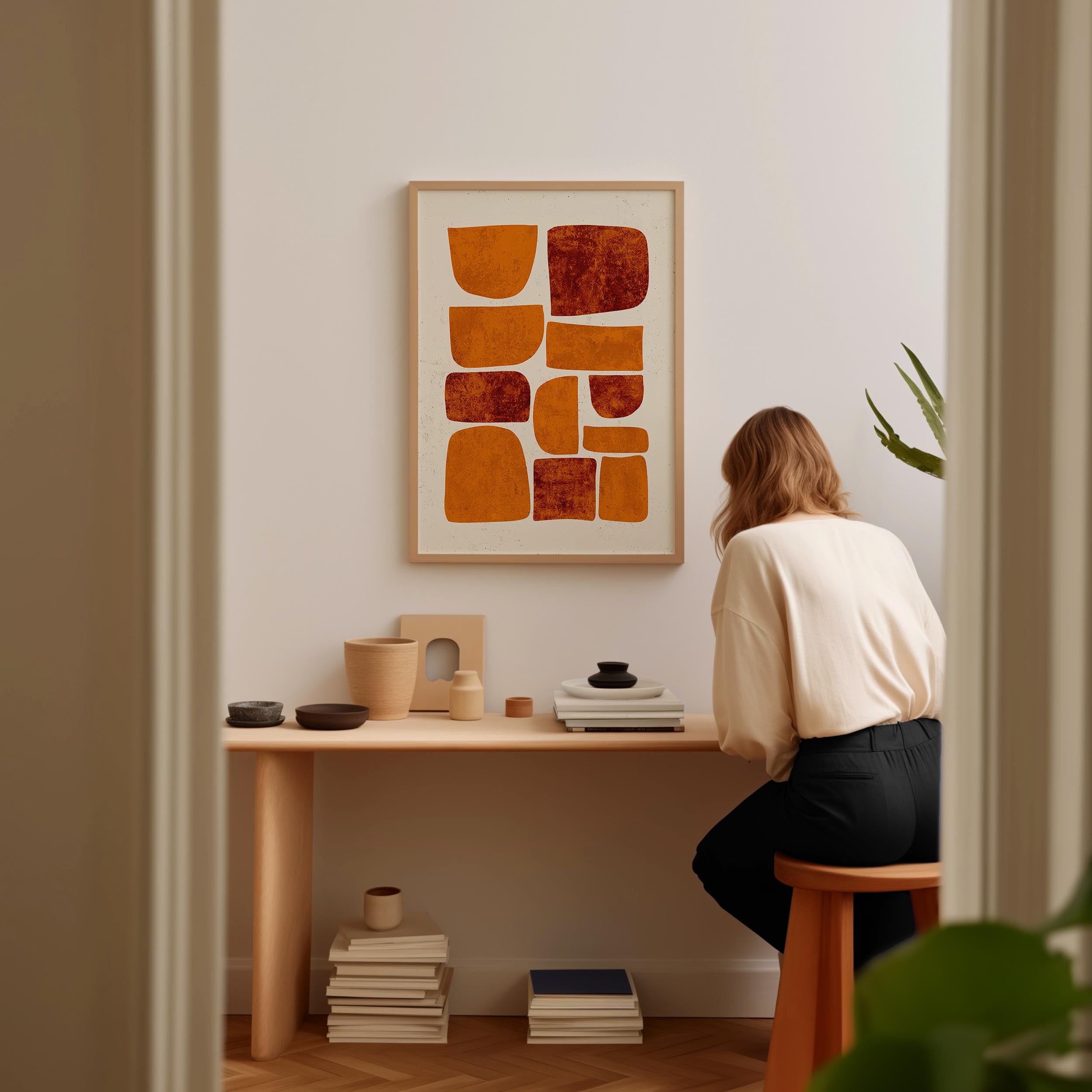 A woman is sitting at a wooden desk, surrounded by various objects, including a framed abstract artwork, a stack of books, and a plant.