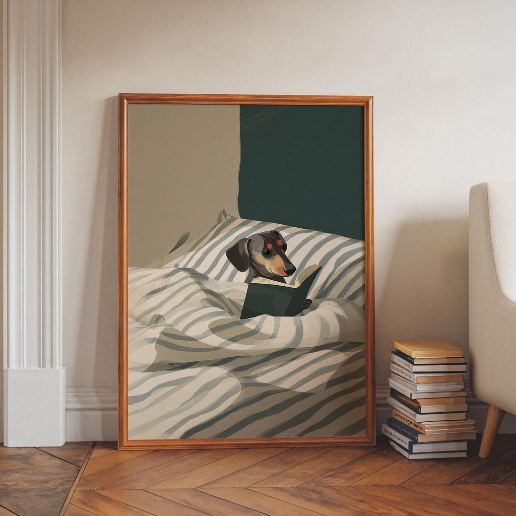 A framed illustration of a dachshund dog lying on a bed and reading a book.