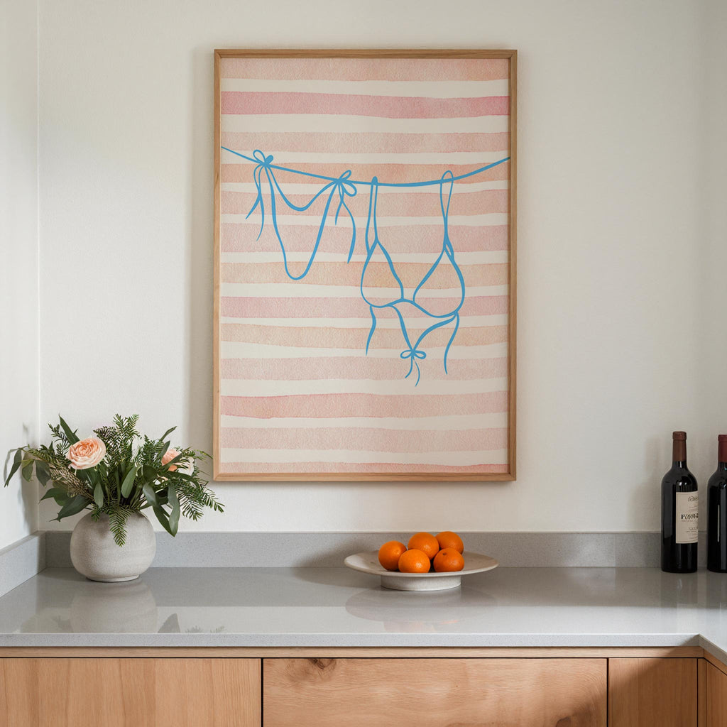 a kitchen counter with a framed artwork of a pink and white striped design hanging above it. On the counter, there is a vase with flowers, a plate of oranges, and two bottles of wine.