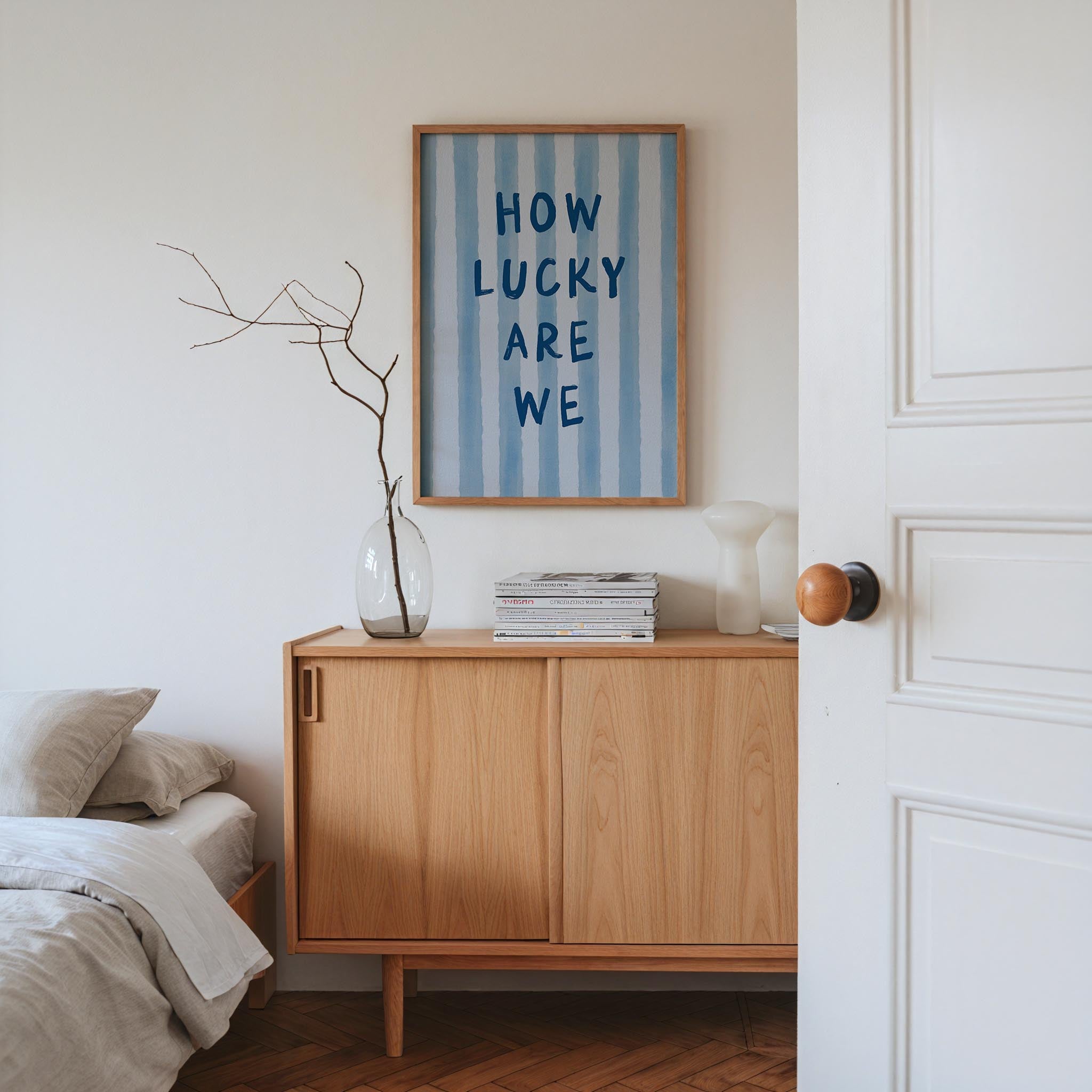 a bedroom with a wooden dresser, a framed poster with the text "HOW LUCKY ARE WE" above it, a vase with branches, and a stack of books on the dresser.
