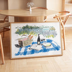A framed illustration depicting a picnic scene with a basket of food, a bottle of wine, and a book on a blue and white checkered blanket, set against a scenic landscape.