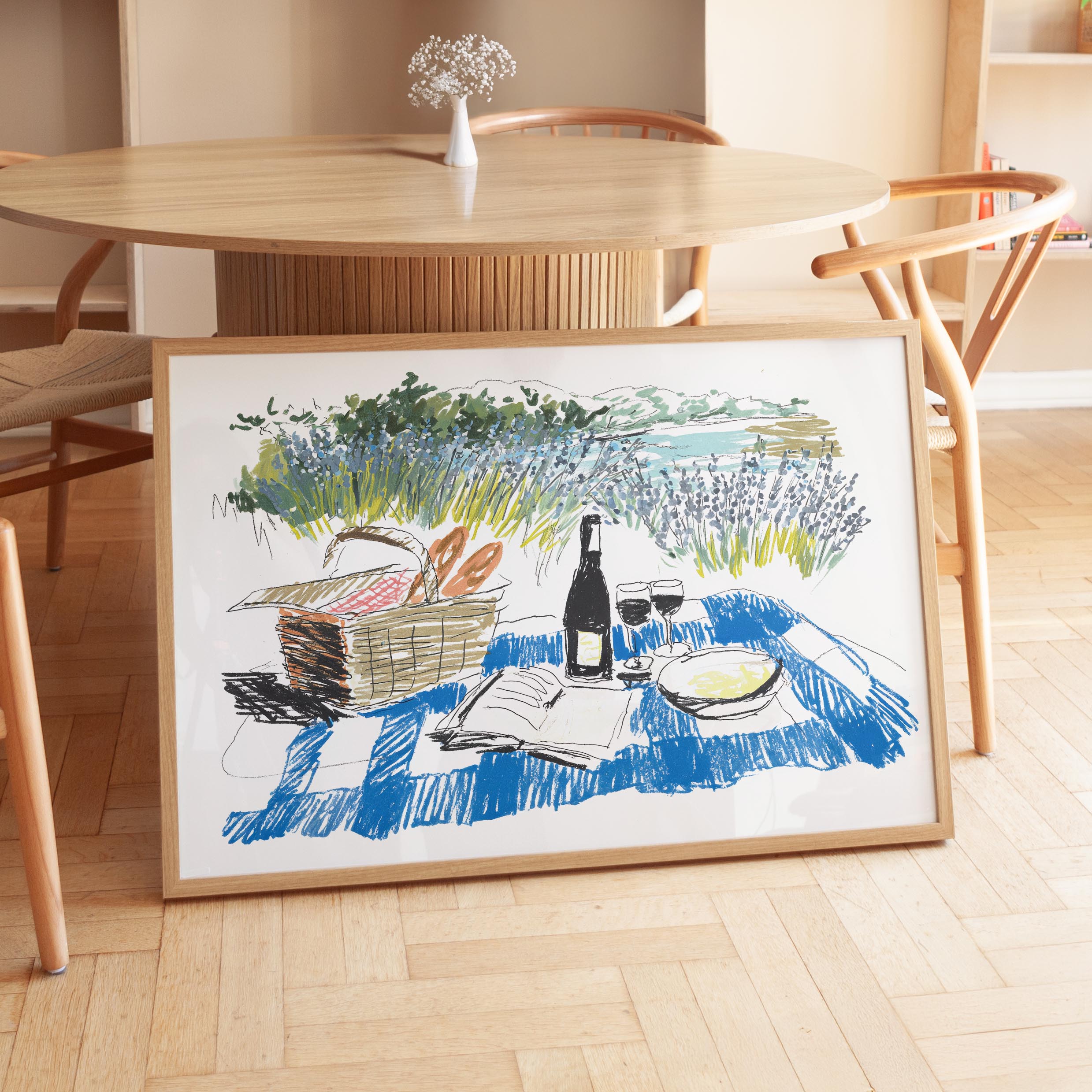 A framed illustration depicting a picnic scene with a basket of food, a bottle of wine, and a book on a blue and white checkered blanket, set against a scenic landscape.