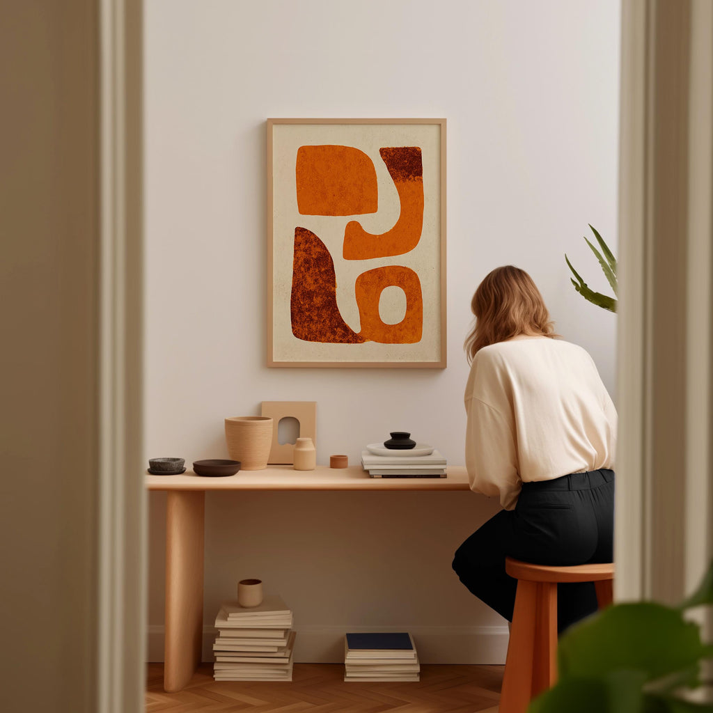 A woman is sitting at a wooden desk, surrounded by various objects, including a framed abstract artwork, a stack of books, and a plant.