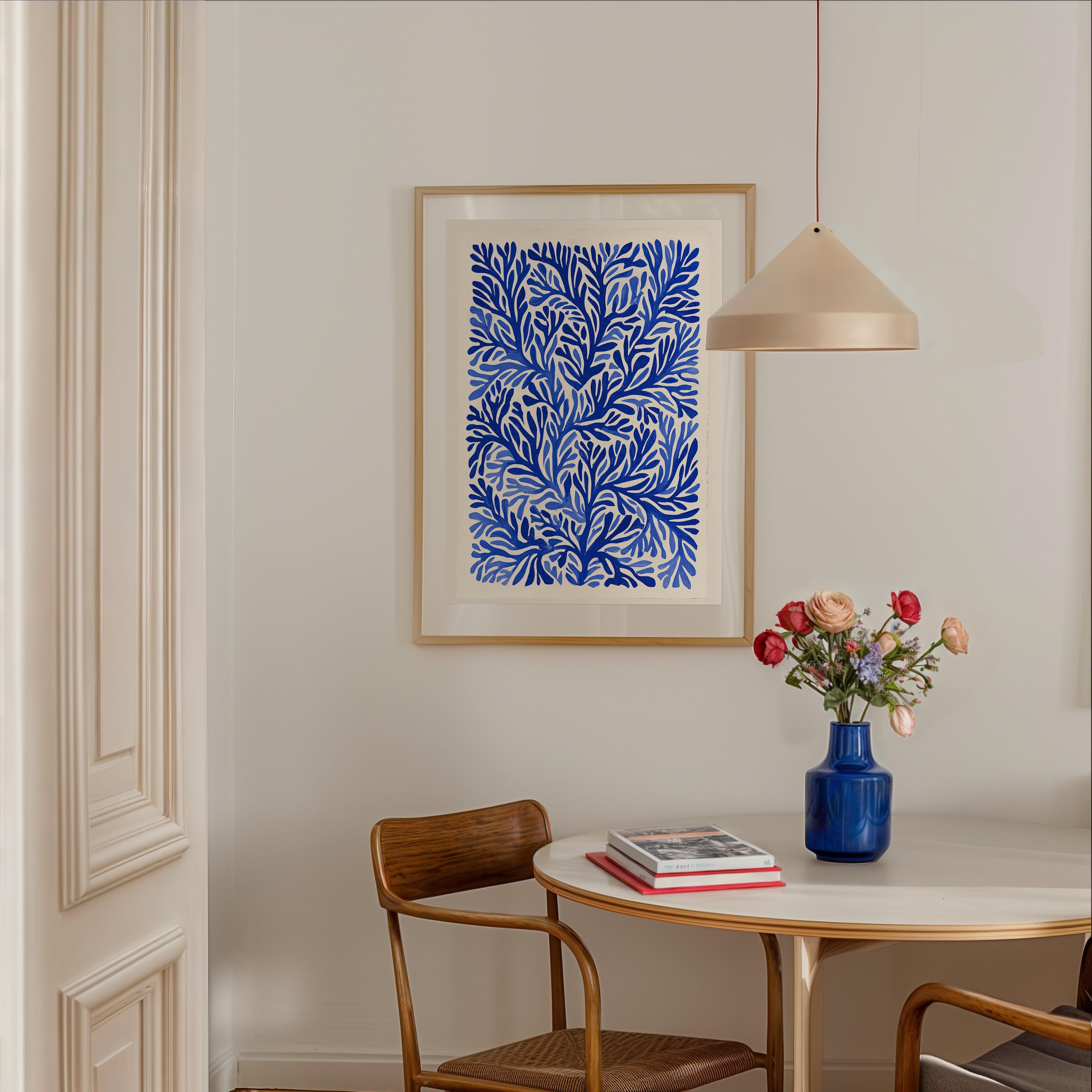a dining area with a white table, a blue vase with flowers, and a framed print of blue and white floral designs hanging on the wall above the table.