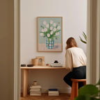A woman is sitting at a wooden desk, looking at a framed painting of a vase with white tulips hanging on the wall above it.