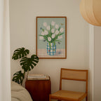 a cozy corner of a room with a framed painting of white tulips in a blue and white vase, a wooden chair, a stack of books, and a potted plant.