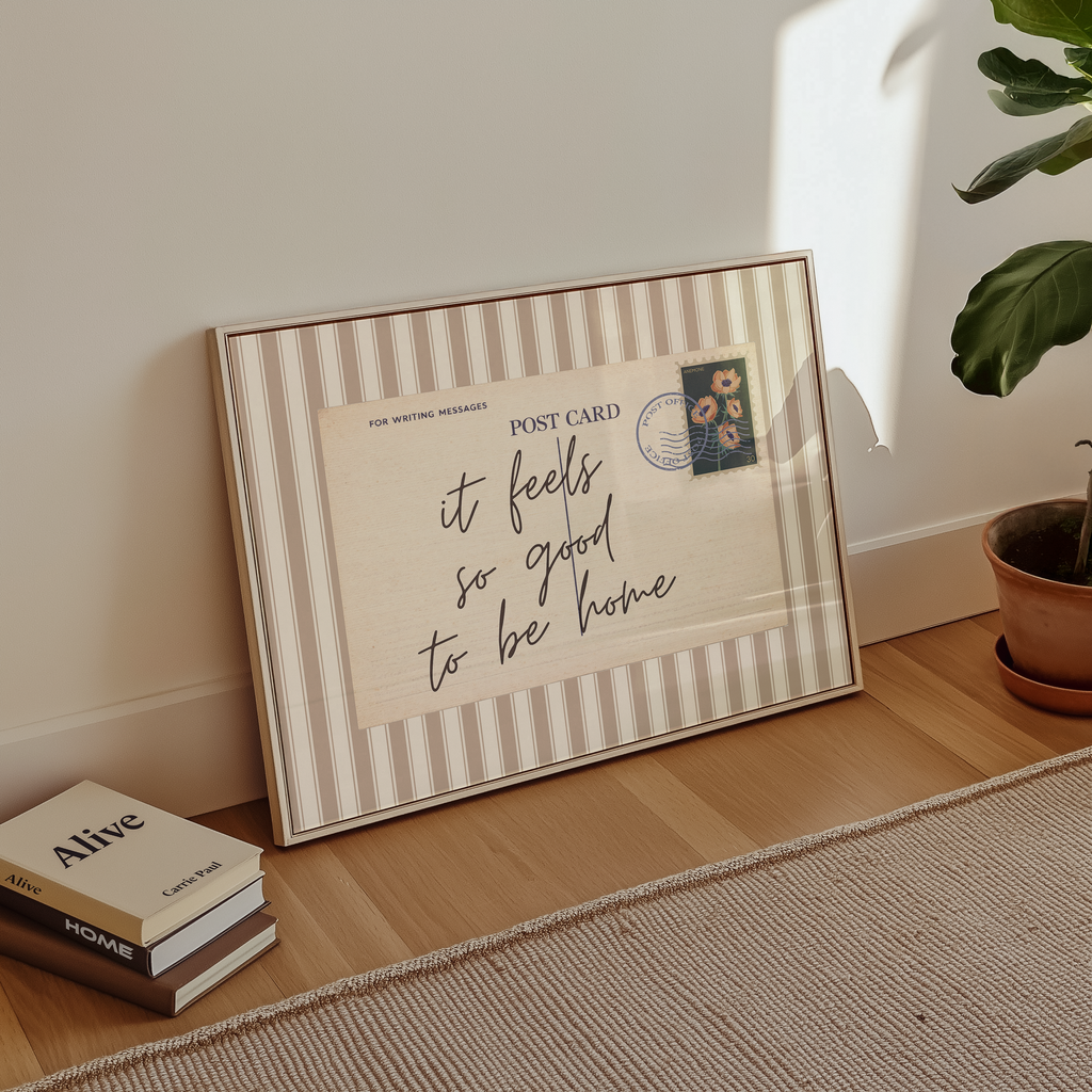 A framed postcard with a handwritten message sits on a wooden floor next to a stack of books and a potted plant.