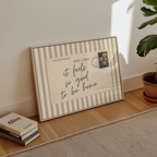 A framed postcard with a handwritten message sits on a wooden floor next to a stack of books and a potted plant.