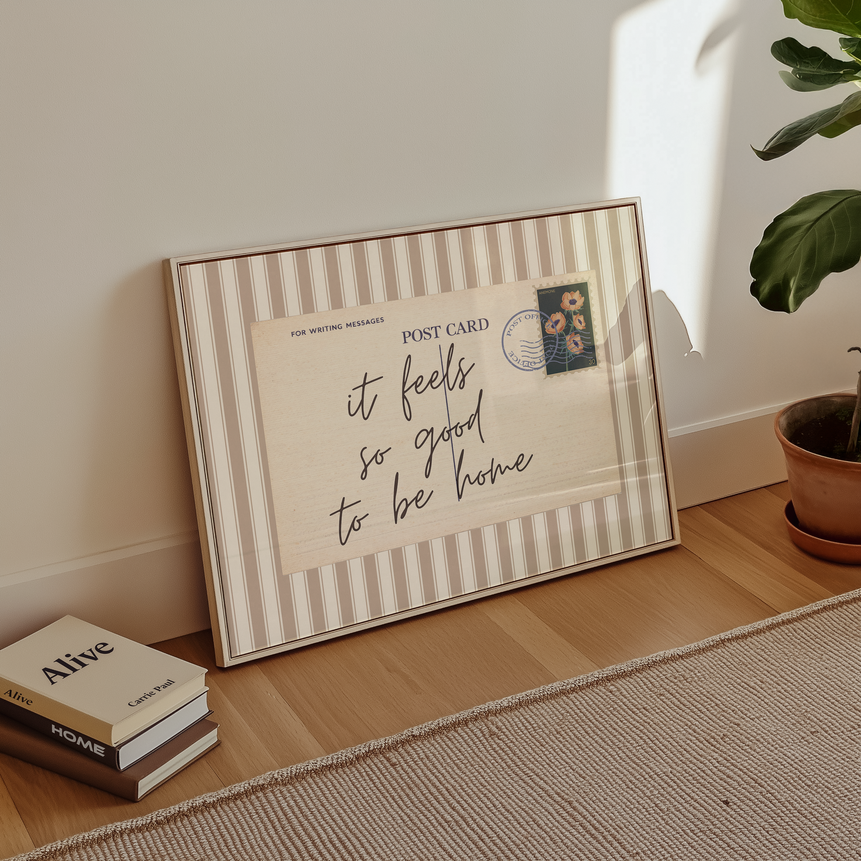 A framed postcard with a handwritten message sits on a wooden floor next to a stack of books and a potted plant.