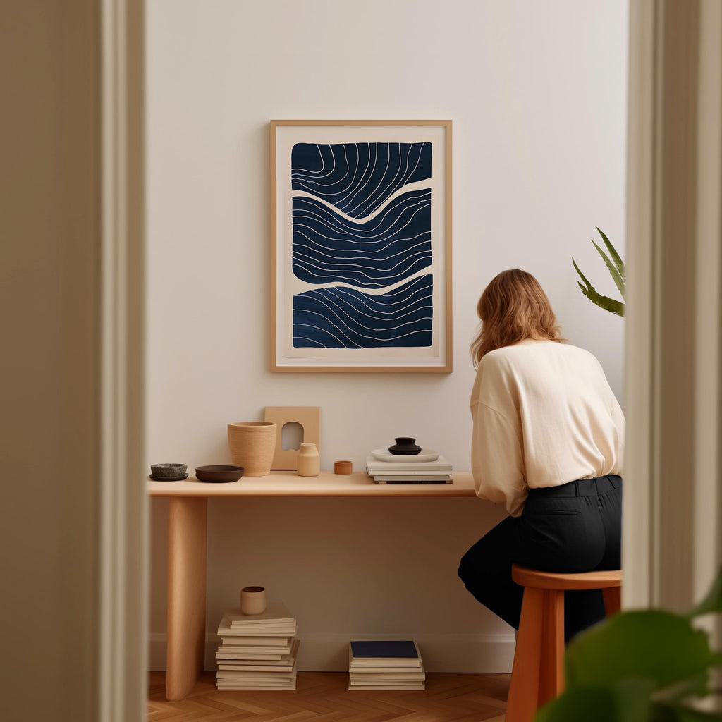 A woman is sitting at a wooden desk, surrounded by various objects, including a framed abstract artwork, a stack of books, and a plant.