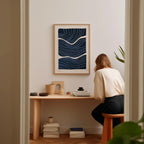 A woman is sitting at a wooden desk, surrounded by various objects, including a framed abstract artwork, a stack of books, and a plant.
