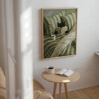 a cozy scene with a framed painting of a cat sitting on a bed, reading a book, and enjoying a cup of coffee. The painting is hung on a white wall above a small wooden table with a book and a cup of coffee on it.