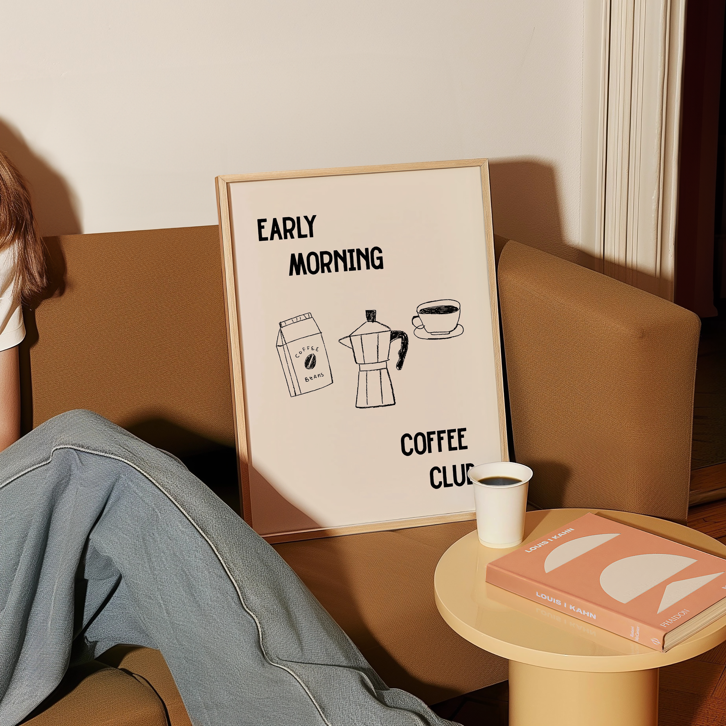 a person sitting on a couch with a framed poster that reads "Early Morning Coffee Club" in front of them.