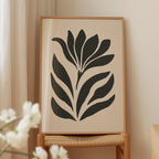 a framed artwork of a stylized flower or plant with black and white elements, displayed on a wooden chair against a light-colored wall.