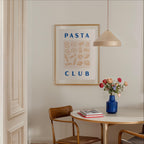 a dining area with a table, chairs, and a framed poster on the wall. The poster displays the text "PASTA CLUB" and includes illustrations of various pasta shapes. A vase of flowers is placed on the table, and there are books and a lamp in the room.