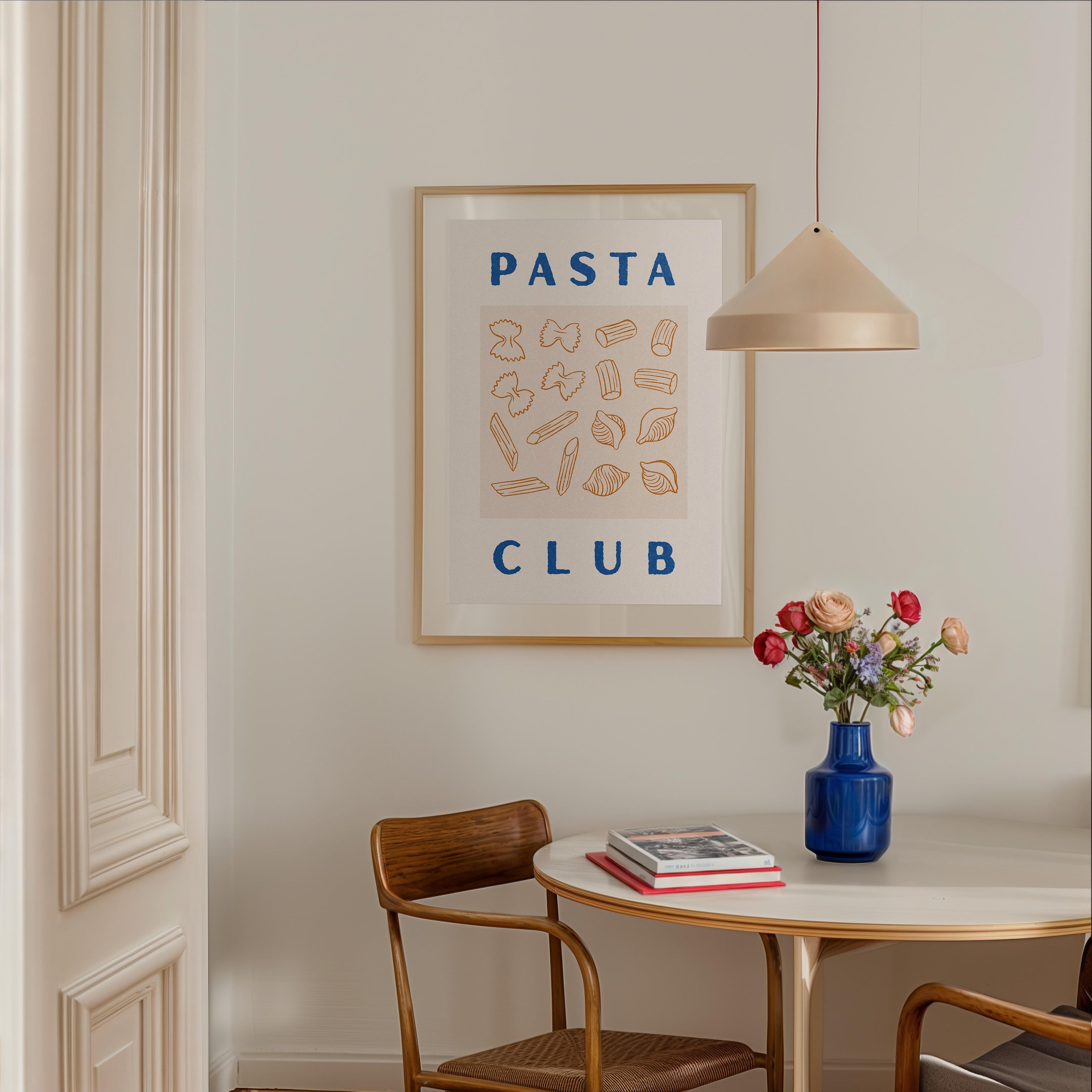a dining area with a table, chairs, and a framed poster on the wall. The poster displays the text "PASTA CLUB" and includes illustrations of various pasta shapes. A vase of flowers is placed on the table, and there are books and a lamp in the room.