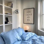 a cozy bedroom with a bed, a bookshelf, and a framed poster on the wall. The poster features the phrase "How Lucky Are We" in bold letters.