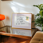 a framed postcard with a handwritten message, placed on a wooden table next to a potted plant.