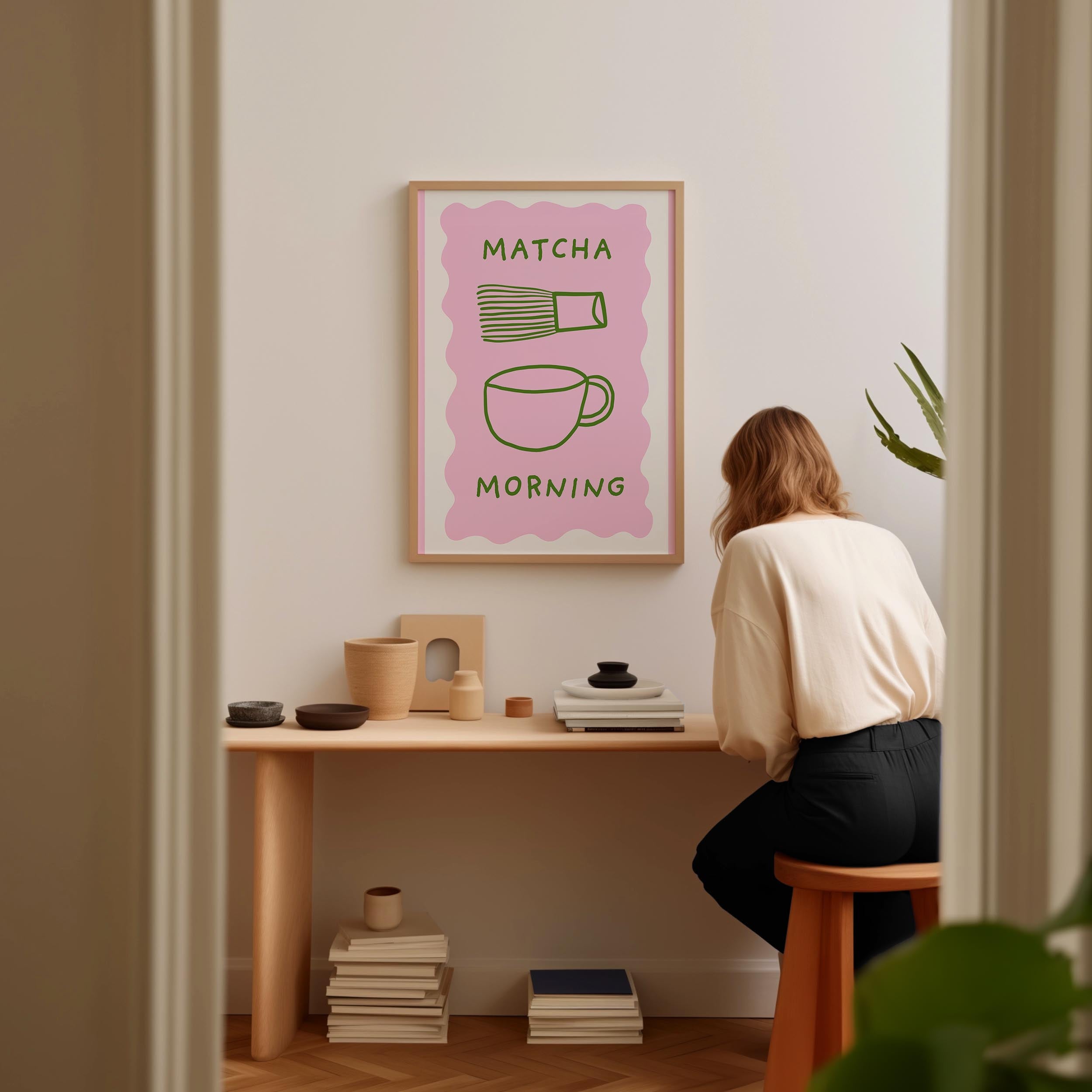 A woman is sitting at a wooden desk, surrounded by various objects, including a framed poster with the text "MATCHA MORNING" and illustrations of a cup and a bowl.