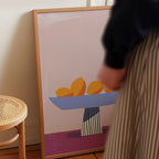 a person standing next to a framed artwork depicting a bowl of fruit on a table.