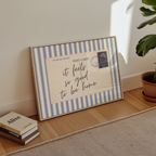 A framed postcard with a handwritten message sits on a wooden floor next to a stack of books and a potted plant.