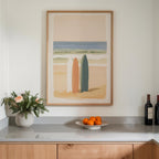 a framed painting of two surfboards on a wall above a counter with a vase of flowers and a plate of oranges.