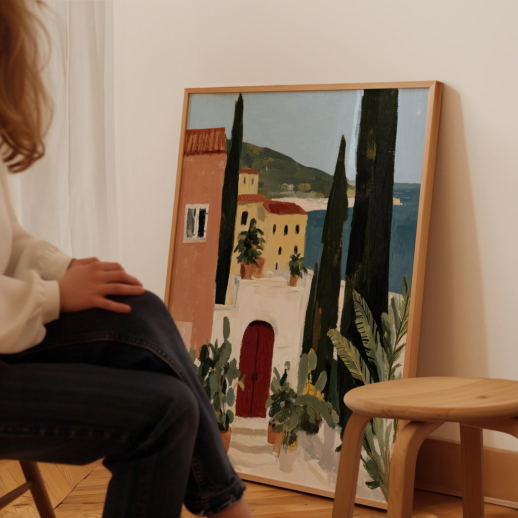 A person is sitting in front of a large painting depicting a coastal town with buildings, trees, and a red door.