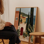 A person is sitting in front of a large painting depicting a coastal town with buildings, trees, and a red door.