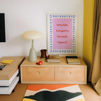 a wooden dresser with a lamp, a framed poster, and a stack of books on top. The dresser has two drawers and a shelf. The poster on the wall has the words "sunrise, sunset, repeat" written on it in a colorful, artistic style.