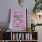 a pink poster with the text "MATCHA MORNING" displayed on it, placed on a bookshelf next to a lamp and other decorative items.