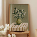 a framed painting of a vase with green leaves on a striped tablecloth.