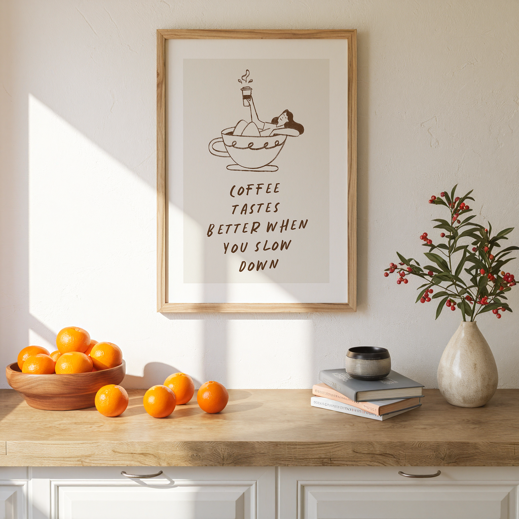 a framed poster with a cartoon illustration of a person drinking coffee, accompanied by the text "Coffee tastes better when you slow down". The poster is displayed on a wall above a wooden table with a bowl of oranges, a vase of flowers, and a stack of books.