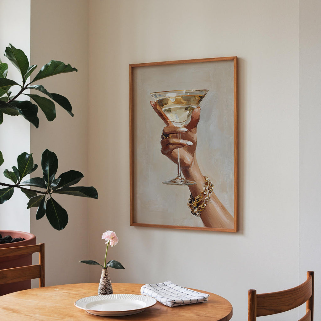 a framed painting of a hand holding a martini glass, with a vase of pink flowers and a plate of food on a wooden table in the foreground.