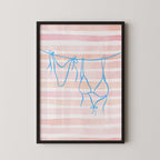 A framed print with a pink and white striped background and blue bra straps hanging from a line.