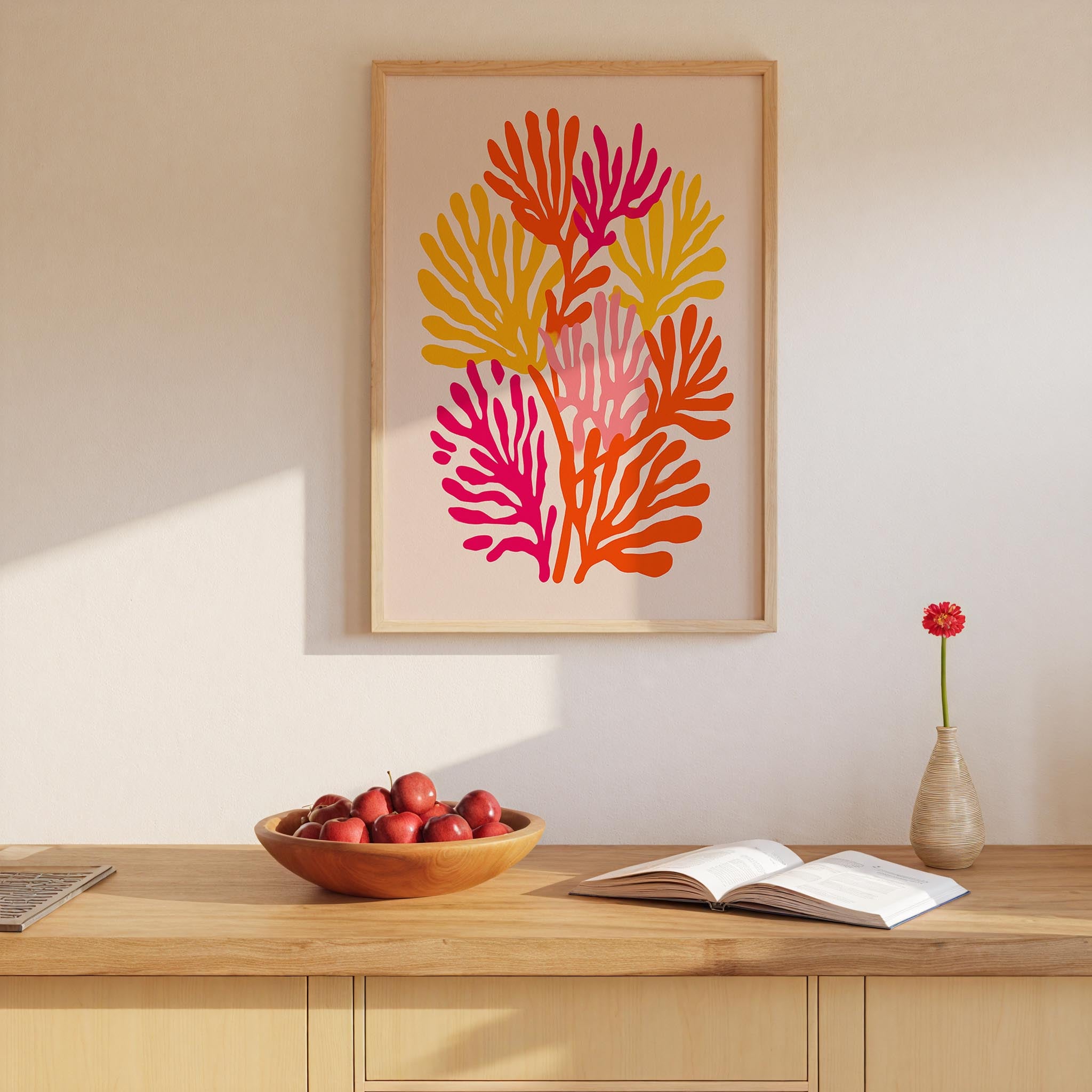 a wooden table with a bowl of red apples, an open book, and a vase with a single red flower. Above the table, there is a framed artwork depicting a colorful abstract design with orange, pink, and yellow elements.