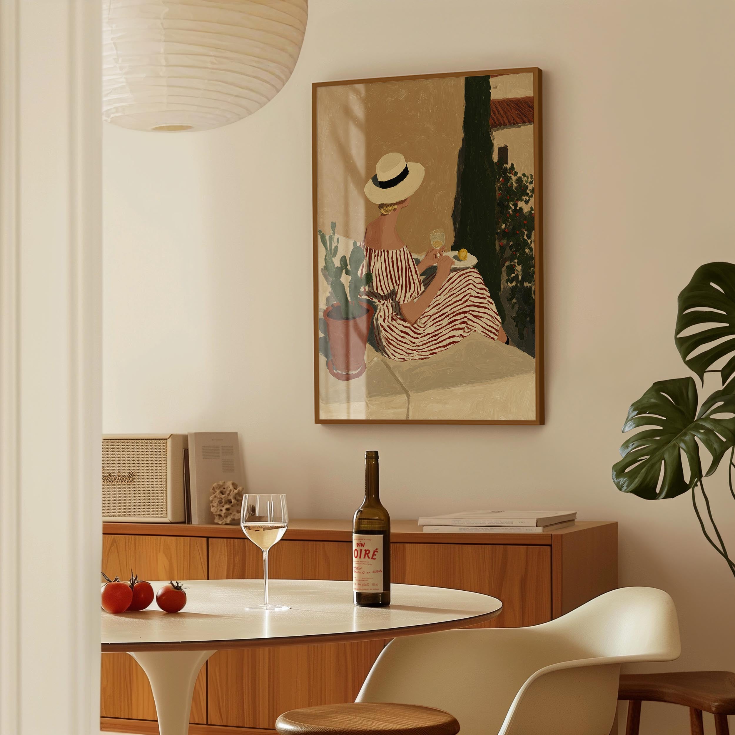 a dining area with a table, chairs, and a framed painting of a woman in a striped dress. There is a bottle of wine and a glass of wine on the table, along with some tomatoes. The painting is hanging on the wall above the table.