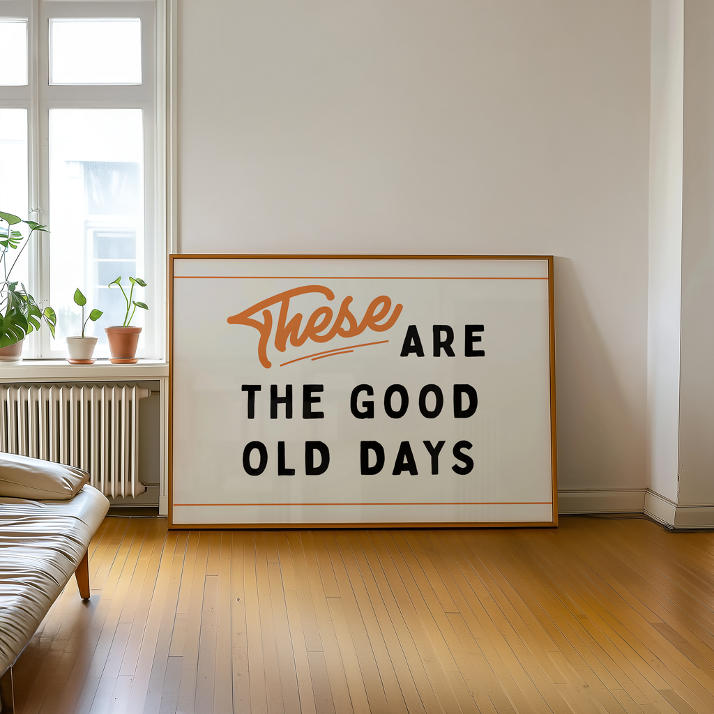 a large framed sign with the text "These are the good old days" on it, placed on a wooden floor in a room with a window and a plant.