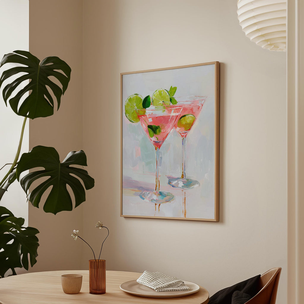 a framed painting of two martini glasses with a pink drink and lime slices, hanging on a wall above a wooden table with a vase of flowers and a plate of food.