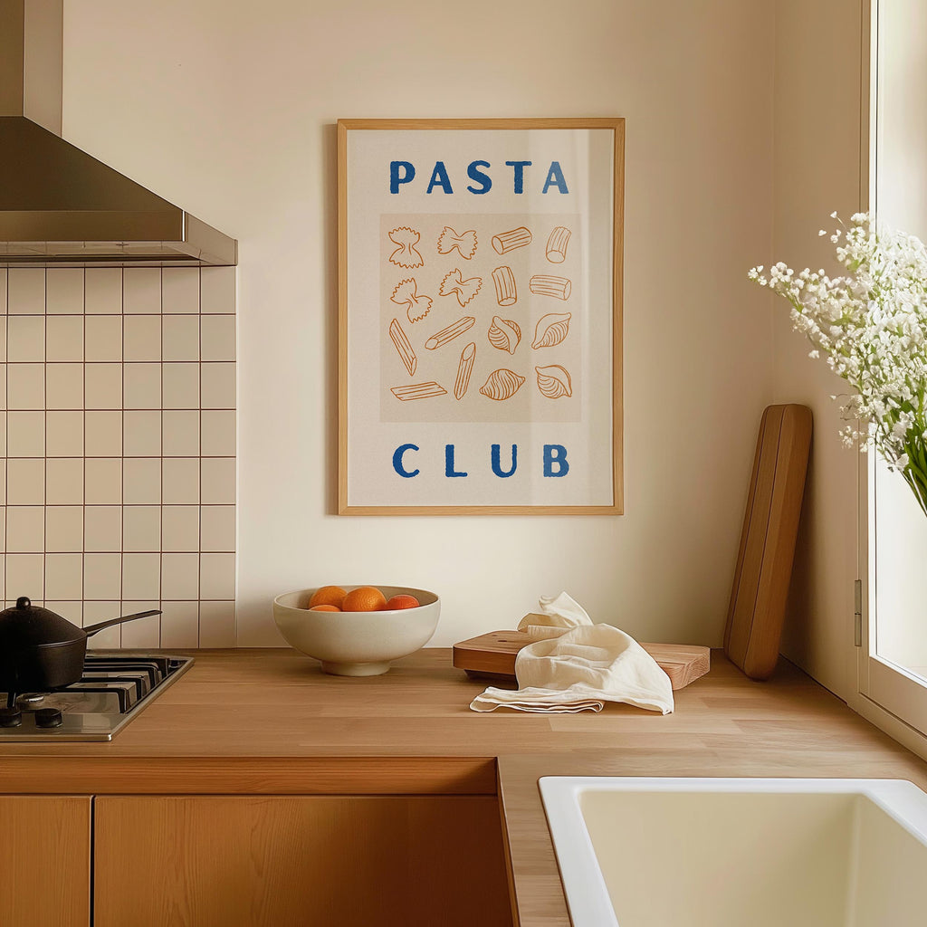 a kitchen counter with a framed poster of pasta and a bowl of fruit, including oranges and a vase of white flowers.