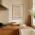 a kitchen counter with a framed poster of pasta and a bowl of fruit, including oranges and a vase of white flowers.